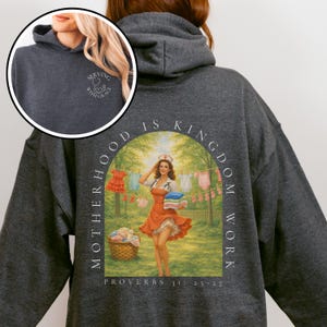 Op de afbeelding: Donkergrijze hoodie met een kleine witte afbeelding op de voorkant en een grote, kleurrijke illustratie op de achterkant. De afbeelding op de achterkant toont een vrouw in een verpleegstersuniform en de tekst "MOTHERHOOD IS KINGDOM WORK".