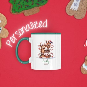 May include: A white ceramic mug with a green rim features a festive Christmas-themed design. The letter "E" is decorated with lights, candy canes, and a reindeer. The mug is personalized with the name "Emily" and the year "2023".