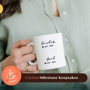 Könnte beinhalten: Weiße Keramiktasse mit schwarzem Text, der "Schwester ♥ EST. 2000" und "Tante ♥ EST. 2024" lautet.