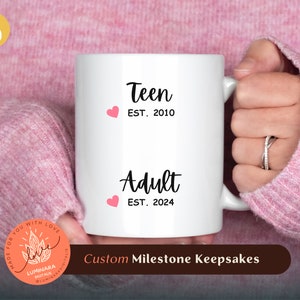 Könnte beinhalten: Eine weiße Keramiktasse mit schwarzem Text, der "Teen EST. 2010" und "Adult EST. 2024" lautet. Die Tasse hat zwei rosa Herzen darauf. Die Tasse wird von einer Hand gehalten, die einen rosa Pullover trägt. Der Text "Custom Milestone Keepsakes" befindet sich am unteren Rand des Bildes.