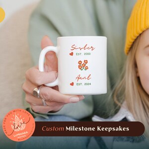Könnte beinhalten: Eine weiße Keramiktasse mit dem Text "Sister Est. 2000" und "Aunt Est. 2024" mit einem roten Herzen und roten Blumen dazwischen. Die Tasse wird von einer Hand gehalten. Der Text "Custom Milestone Keepsakes" ist am unteren Rand des Bildes sichtbar.