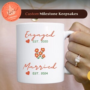 Könnte beinhalten: Eine weiße Keramiktasse mit dem Text "Engaged EST. 2020" und "Married EST. 2024" in schwarzer Kursivschrift. Die Tasse hat ein rotes Herz und eine Gruppe roter Blumen zwischen den beiden Textzeilen.