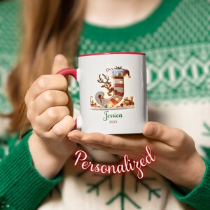 Peut inclure: Mug en céramique blanche avec un bord rouge. Le mug représente une chaussette de Noël avec un renne et la lettre "J". Le mug est personnalisé avec le nom "Jessica" et l'année "2023".