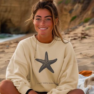 May include: A beige sweatshirt featuring a dark gray starfish graphic. The sweatshirt has long sleeves and the text "SIMPLY SANTA BARBARA" printed below the starfish. The person is sitting on a sandy beach.