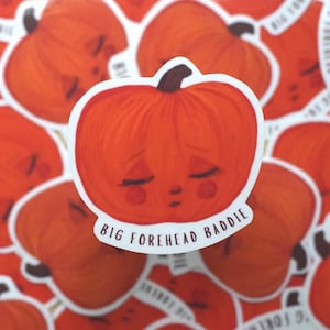 May include: A collection of orange pumpkin stickers with a sad face and the text "BIG FOREHEAD BADDIE". The stickers are a vibrant orange color with a white border and a black stem.