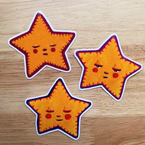 May include: Three yellow star-shaped stickers with embroidered edges and friendly faces. Each star has a different facial expression.