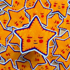 May include: A close-up of a pile of yellow star-shaped stickers with a smiling face and closed eyes. The stars have a blue outline and red cheeks.