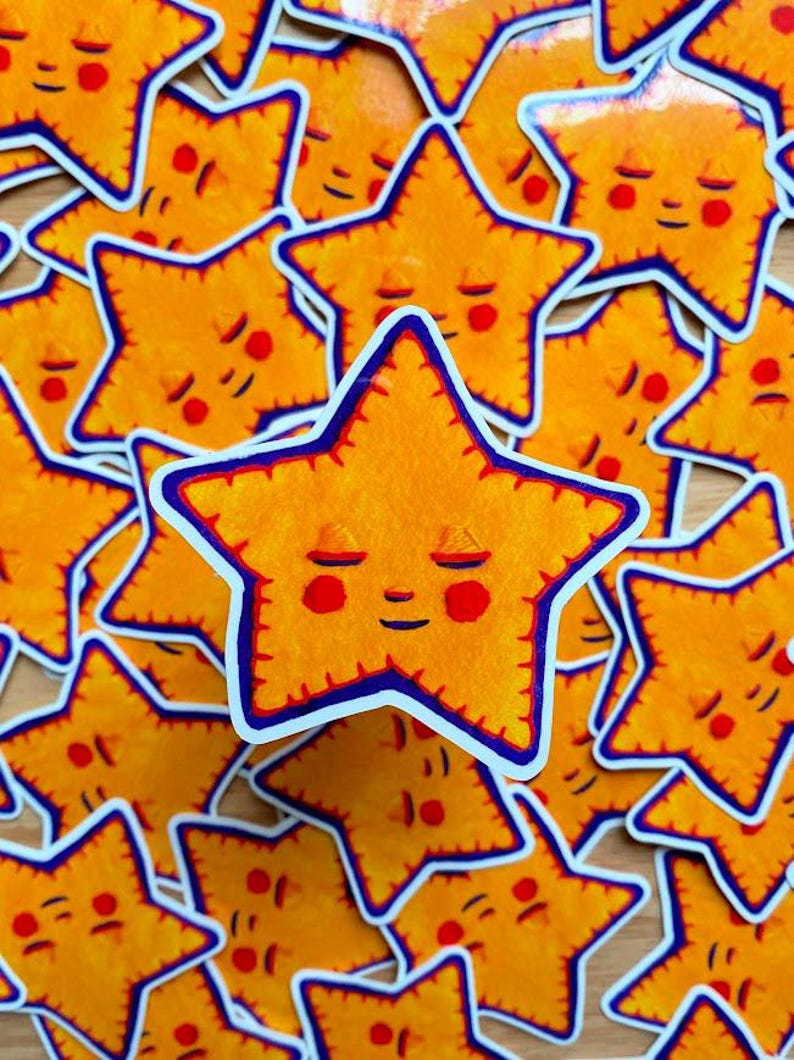 May include: A close-up of a pile of yellow star-shaped stickers with a smiling face and closed eyes. The stars have a blue outline and red cheeks.