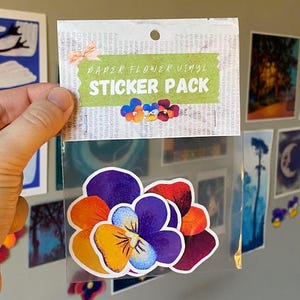 May include: A clear plastic package containing a sticker pack. The sticker pack has a green banner that reads "PAPER FLOWER VINYL STICKER PACK". The stickers are colorful flower designs in shades of purple, orange, and red.