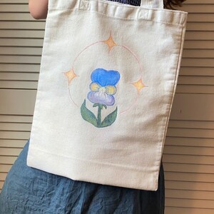 Peut inclure: Un sac fourre-tout en toile blanche avec un motif peint &agrave; la main d'une pens&eacute;e bleue et jaune avec des feuilles vertes. La fleur est entour&eacute;e d'un cercle rose avec quatre &eacute;toiles jaunes.