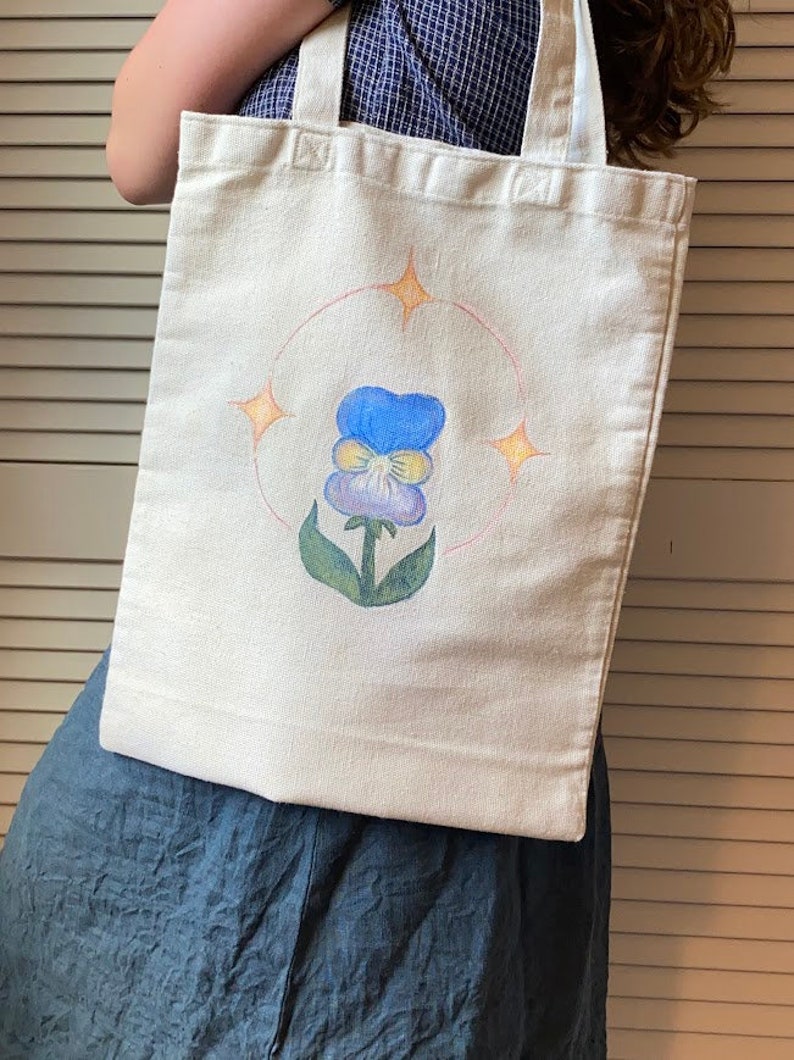 Peut inclure: Un sac fourre-tout en toile blanche avec un motif peint &agrave; la main d'une pens&eacute;e bleue et jaune avec des feuilles vertes. La fleur est entour&eacute;e d'un cercle rose avec quatre &eacute;toiles jaunes.