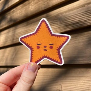 May include: A sticker of a cartoon orange starfish with a stitched border and a sleepy face. The starfish is outlined in maroon.
