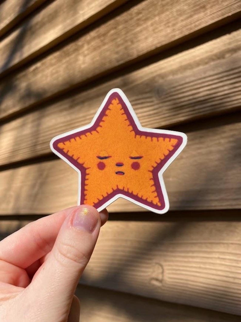 May include: A sticker of a cartoon orange starfish with a stitched border and a sleepy face. The starfish is outlined in maroon.