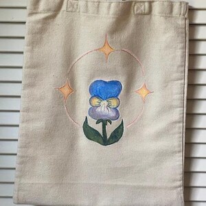 Peut inclure: Un sac fourre-tout en toile blanche avec un motif peint &agrave; la main d'une pens&eacute;e bleue et jaune avec des feuilles vertes. La fleur est entour&eacute;e d'un cercle avec trois &eacute;toiles jaunes.