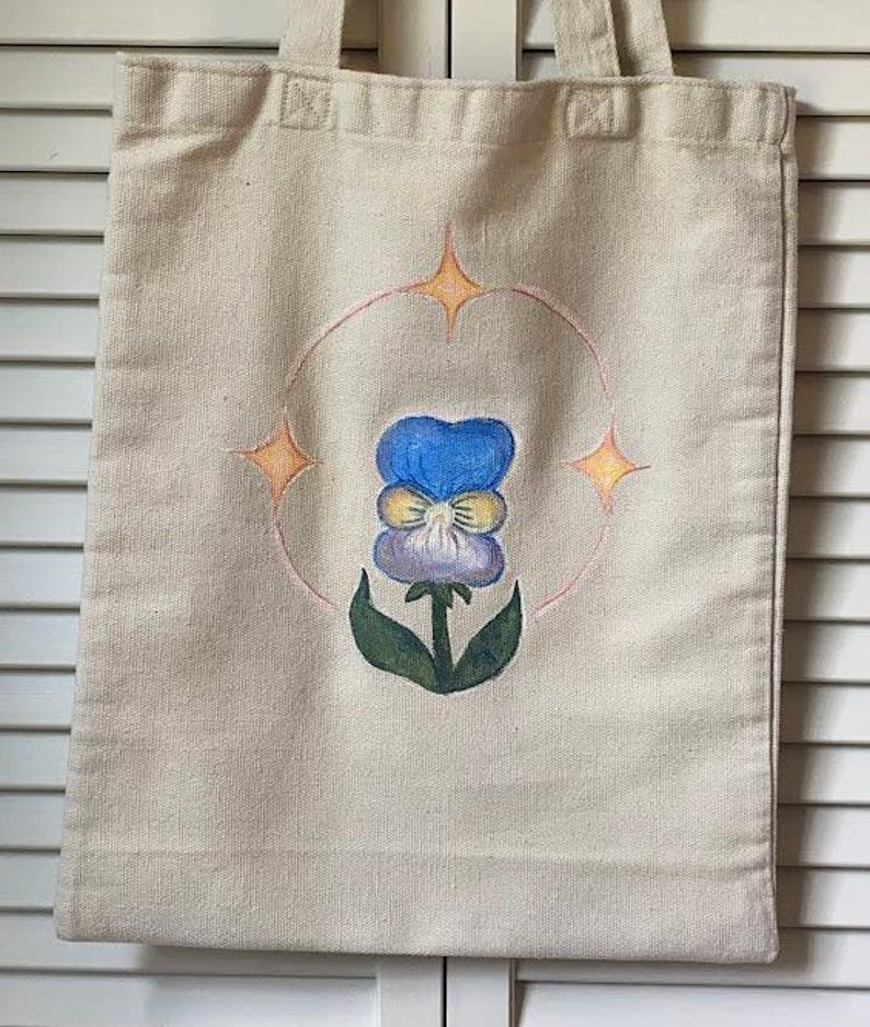 Peut inclure: Un sac fourre-tout en toile blanche avec un motif peint &agrave; la main d'une pens&eacute;e bleue et jaune avec des feuilles vertes. La fleur est entour&eacute;e d'un cercle avec trois &eacute;toiles jaunes.