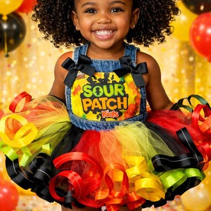 Puede incluir: Una niña pequeña con un tutú colorido y una parte superior de mezclilla con el texto "SOUR PATCH Kids". El tutú presenta capas de cintas rojas, naranjas, amarillas, verdes y negras. Lazos negros adornan la parte superior y el cabello.