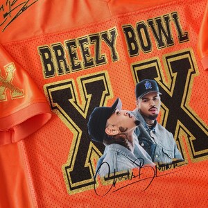Puede incluir: Camiseta de malla naranja con el texto "BREEZY BOWL" y un gr&aacute;fico de dos personas. La camiseta tiene detalles dorados, estrellas negras y firmas. La camiseta es de cuello en V.