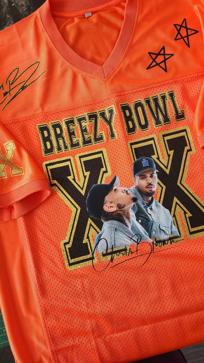Puede incluir: Camiseta de malla naranja con el texto "BREEZY BOWL" y un gr&aacute;fico de dos personas. La camiseta tiene detalles dorados, estrellas negras y firmas. La camiseta es de cuello en V.