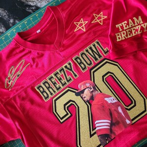Puede incluir: Una camiseta de malla roja con letras y detalles dorados. La camiseta presenta el texto "BREEZY BOWL" y el n&uacute;mero "20" con una imagen de una persona. Las mangas tienen impreso "TEAM BREEZY". Dos estrellas doradas est&aacute;n encima del texto.