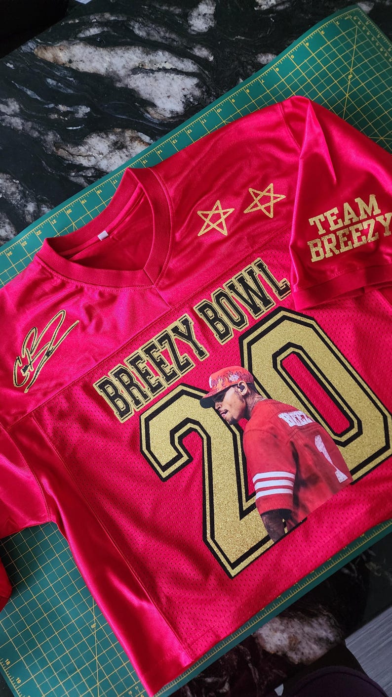 Puede incluir: Una camiseta de malla roja con letras y detalles dorados. La camiseta presenta el texto "BREEZY BOWL" y el n&uacute;mero "20" con una imagen de una persona. Las mangas tienen impreso "TEAM BREEZY". Dos estrellas doradas est&aacute;n encima del texto.