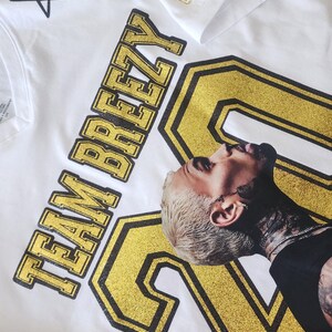 Puede incluir: Camisetas blancas con detalles dorados brillantes. Una camiseta muestra el texto "TEAM BREEZY" y el n&uacute;mero "20" con un retrato. Otra camiseta tiene dise&ntilde;os de "XX" y estrellas doradas brillantes. Las camisetas est&aacute;n sobre una esterilla de corte verde.