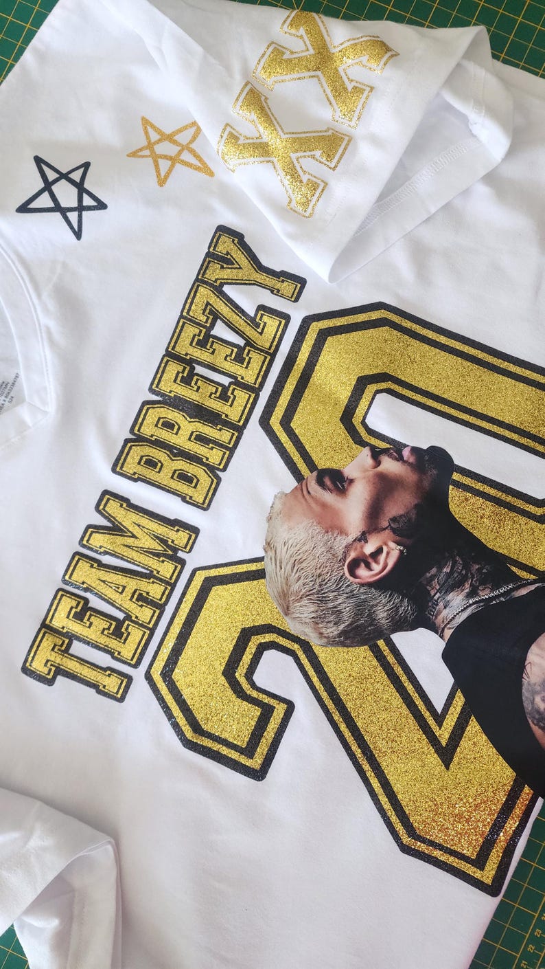 Puede incluir: Camisetas blancas con detalles dorados brillantes. Una camiseta muestra el texto "TEAM BREEZY" y el n&uacute;mero "20" con un retrato. Otra camiseta tiene dise&ntilde;os de "XX" y estrellas doradas brillantes. Las camisetas est&aacute;n sobre una esterilla de corte verde.