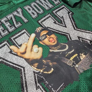 Puede incluir: Camiseta de f&uacute;tbol americano de malla verde con el texto "EEZY BOWL" y una gran "X" en purpurina plateada. La camiseta presenta un gr&aacute;fico de una persona con gafas de sol y una cadena.