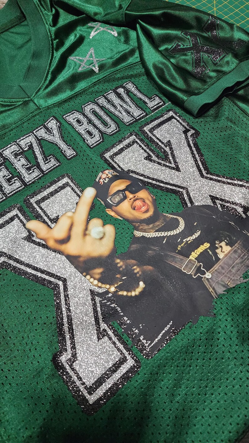 Puede incluir: Camiseta de f&uacute;tbol americano de malla verde con el texto "EEZY BOWL" y una gran "X" en purpurina plateada. La camiseta presenta un gr&aacute;fico de una persona con gafas de sol y una cadena.