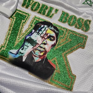 Puede incluir: Camiseta de malla blanca con detalles de purpurina verde y dorada. La camiseta presenta las palabras "WORLD BOSS" y un retrato de una persona con una mano sobre la cara. La manga tiene una hoja de arce dorada.