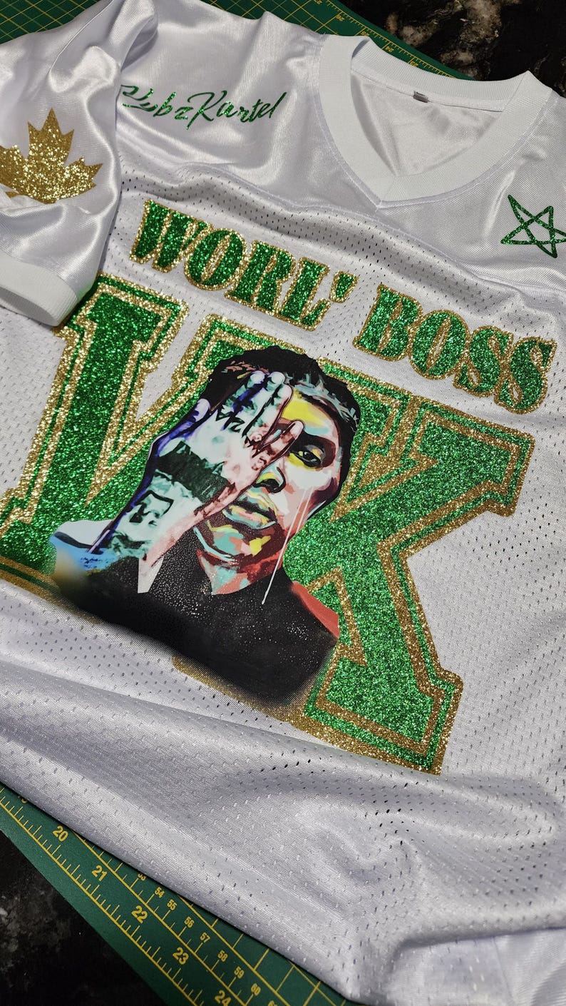 Puede incluir: Camiseta de malla blanca con detalles de purpurina verde y dorada. La camiseta presenta las palabras "WORLD BOSS" y un retrato de una persona con una mano sobre la cara. La manga tiene una hoja de arce dorada.