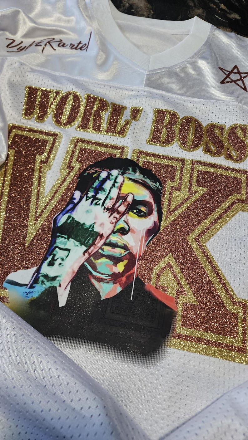 Puede incluir: Camiseta de malla blanca con las palabras "WORK BOSS" en letras doradas con purpurina. Un gr&aacute;fico de una persona cubri&eacute;ndose la cara con una mano est&aacute; en la parte delantera. La camiseta tambi&eacute;n tiene el texto "Zy/Kartel" y una estrella.