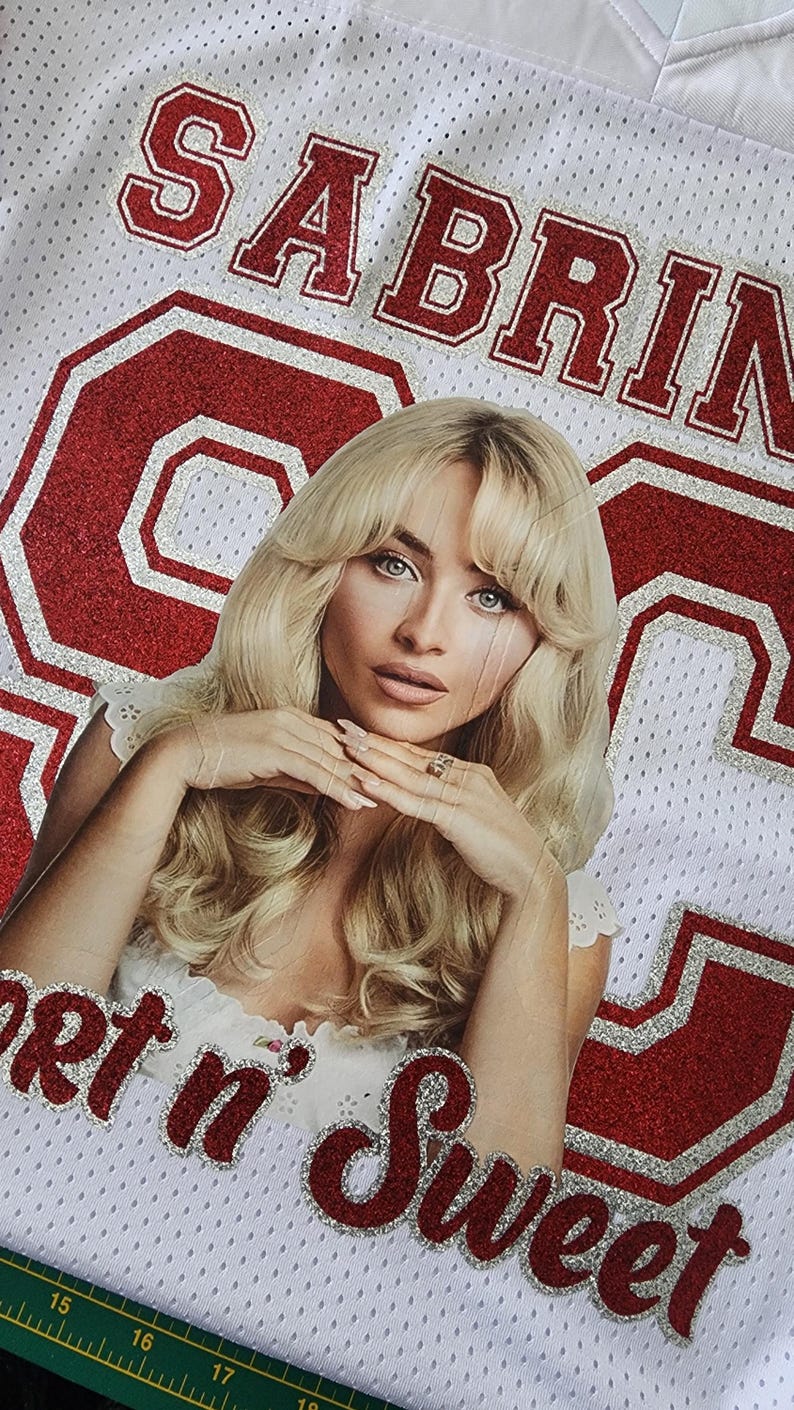 Puede incluir: Camiseta de malla blanca con letras rojas brillantes que dicen "SABRINA" y "Get n' Sweet". Una foto de una mujer con cabello rubio est&aacute; en el centro. El n&uacute;mero 63 tambi&eacute;n est&aacute; en brillo rojo.