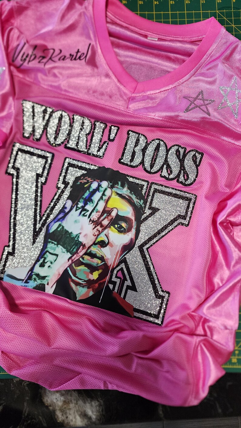 Puede incluir: Camiseta rosa con el texto "WORE' BOSS" y un gr&aacute;fico de la cara de una persona con una mano que la cubre parcialmente. La camiseta tambi&eacute;n tiene el texto "Vybz Kartel" y un dise&ntilde;o de estrella.