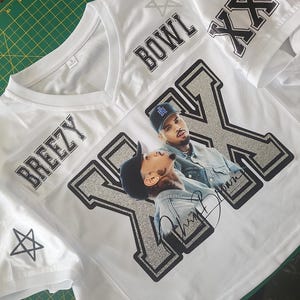 Puede incluir: Camiseta de malla blanca con las palabras "BREEZY" y "BOWL" impresas en negro. La camiseta presenta un gran gr&aacute;fico plateado y negro de una persona, con las letras "XX" en las mangas. Un pentagrama est&aacute; en la manga izquierda.