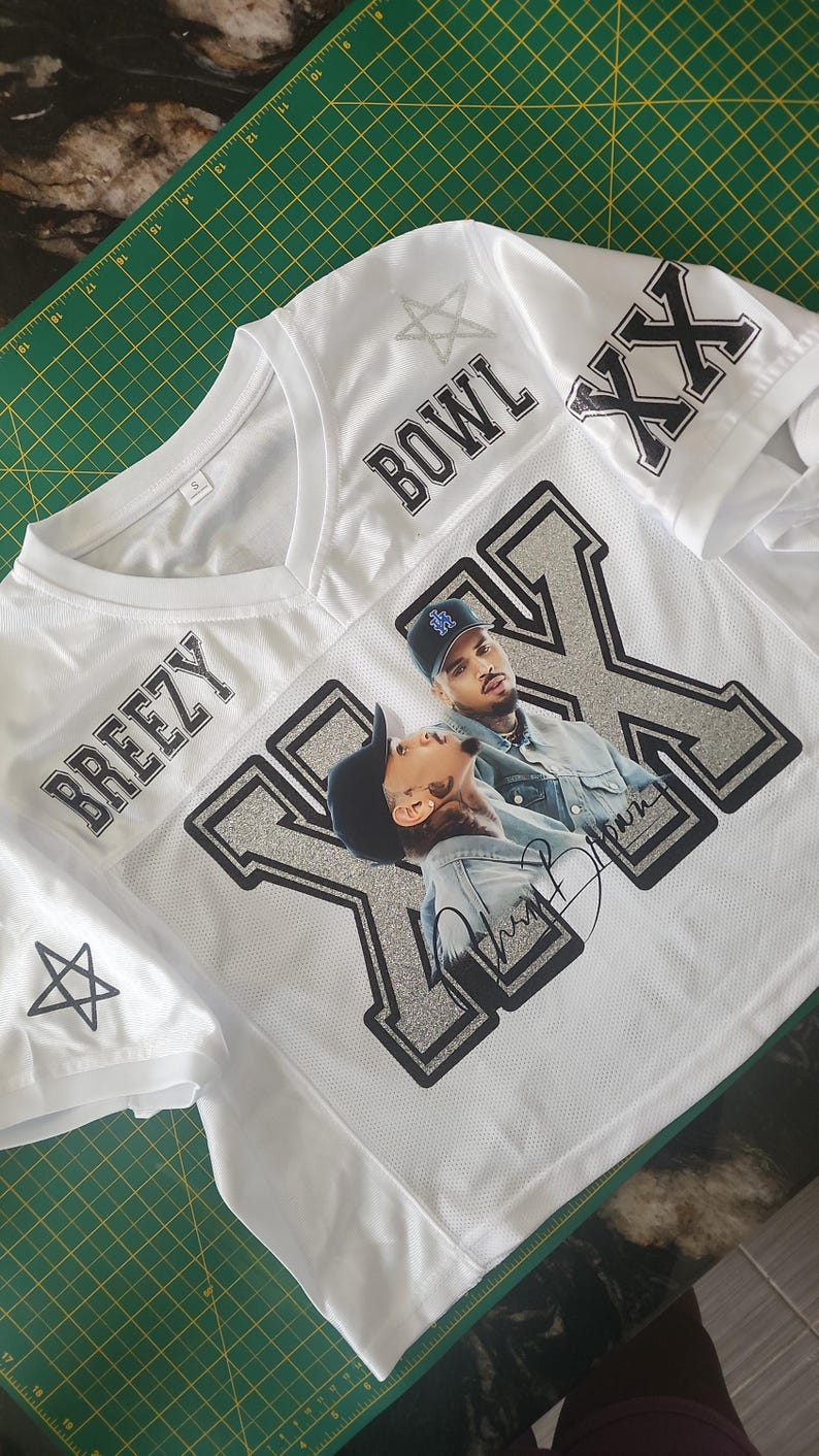 Puede incluir: Camiseta de malla blanca con las palabras "BREEZY" y "BOWL" impresas en negro. La camiseta presenta un gran gr&aacute;fico plateado y negro de una persona, con las letras "XX" en las mangas. Un pentagrama est&aacute; en la manga izquierda.