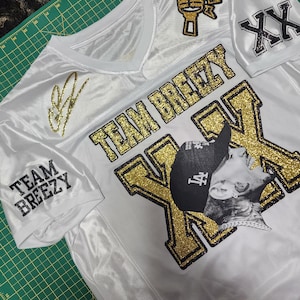 Puede incluir: Jersey blanco con detalles dorados brillantes. Presenta las palabras "TEAM BREEZY" y un gr&aacute;fico de micr&oacute;fono. Incluye una imagen en blanco y negro de una persona con sombrero. La camiseta tambi&eacute;n tiene una firma y las letras "XX".