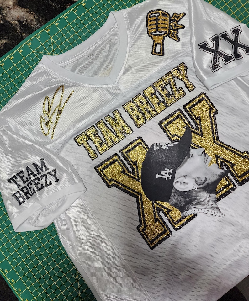 Puede incluir: Jersey blanco con detalles dorados brillantes. Presenta las palabras "TEAM BREEZY" y un gr&aacute;fico de micr&oacute;fono. Incluye una imagen en blanco y negro de una persona con sombrero. La camiseta tambi&eacute;n tiene una firma y las letras "XX".