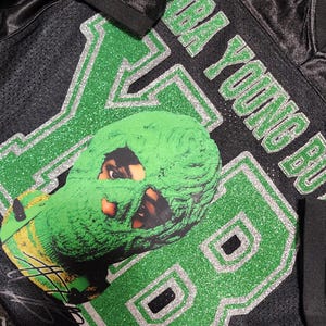 Puede incluir: Camiseta de malla negra con detalles de purpurina verde y plateada. La camiseta lleva el n&uacute;mero "88" y el nombre "NBA YOUNG BOY". Una m&aacute;scara verde con una cara est&aacute; en la parte delantera.