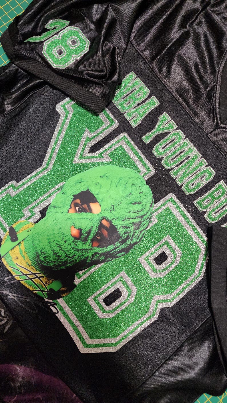 Puede incluir: Camiseta de malla negra con detalles de purpurina verde y plateada. La camiseta lleva el n&uacute;mero "88" y el nombre "NBA YOUNG BOY". Una m&aacute;scara verde con una cara est&aacute; en la parte delantera.