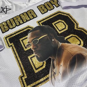 Puede incluir: Camiseta blanca con detalles dorados y negros, con un retrato de una persona con gafas de sol. La camiseta tiene el texto "BURNA BOY" y "TORONTO 2025". Tambi&eacute;n hay estrellas decorativas.