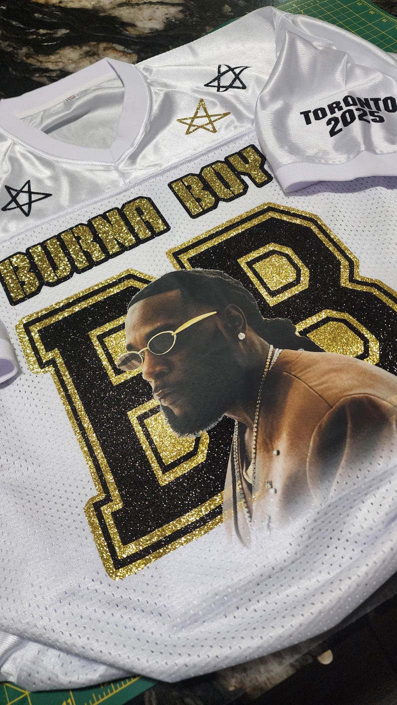 Puede incluir: Camiseta blanca con detalles dorados y negros, con un retrato de una persona con gafas de sol. La camiseta tiene el texto "BURNA BOY" y "TORONTO 2025". Tambi&eacute;n hay estrellas decorativas.