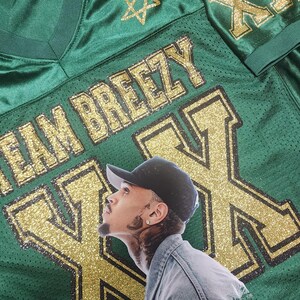 Puede incluir: Camiseta de malla verde con detalles dorados brillantes. La camiseta presenta el texto "TEAM BREEZY" y grandes "XX" doradas con purpurina en la parte delantera. Un retrato de una persona con una gorra negra est&aacute; en la camiseta. Dos estrellas doradas con purpurina est&aacute;n en el hombro.