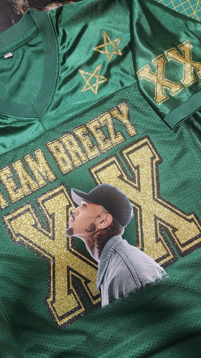 Puede incluir: Camiseta de malla verde con detalles dorados brillantes. La camiseta presenta el texto "TEAM BREEZY" y grandes "XX" doradas con purpurina en la parte delantera. Un retrato de una persona con una gorra negra est&aacute; en la camiseta. Dos estrellas doradas con purpurina est&aacute;n en el hombro.