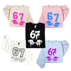 May include: A collection of sweatshirts in various colors, including beige, pink, black, white, and light blue. Each sweatshirt features the number "67" and a hand graphic. The designs are in different colors, such as pink, blue, and white.