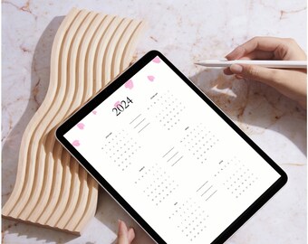 Peut inclure: Une tablette blanche avec un calendrier 2024 affiché à l'écran. La tablette est posée sur une surface en bois et une main tient un stylet.