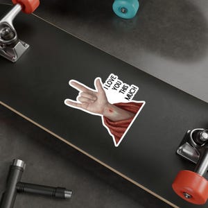 May include: A white sticker with a hand gesture and the text "I LOVE YOU THIS MUCH." The hand is making a rock and roll sign, with a red wound on the arm. The sticker has a red and white border.