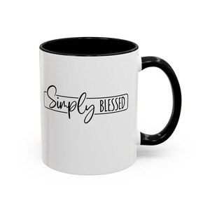 May include: A white ceramic mug with a black interior and a black handle. The exterior features the black text Simply Blessed enclosed within a thin black rectangular outline. The design is centered on the front of the mug.
