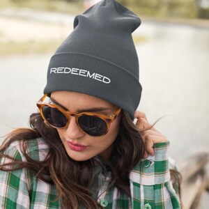 Puede incluir: Un gorro gris oscuro con la palabra "REDEEMED" en letras blancas. La persona lleva gafas de sol carey y una camisa a cuadros verdes y blancos.
