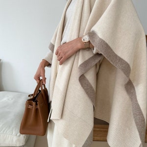 May include: A cream-colored knitted poncho with brown trim. The poncho is draped over a person's shoulders and is worn with cream-colored pants.