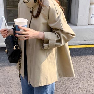 May include: A tan jacket with a high collar and button closure. The jacket has long sleeves with buttoned cuffs and a zipper closure. The person is wearing blue jeans and holding a black purse with a gold chain strap and a white coffee cup.